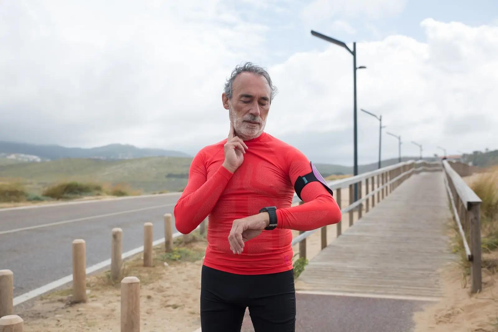 An older man in red running gear checking his watch on a coastal path